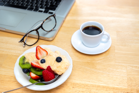 Breakfast bowl of fresh fruits salad with yogurt and coffee and laptop on tableの写真素材