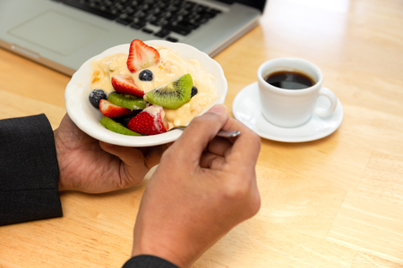 Businessman having bowl of fresh fruits salad with yogurt and coffee and laptop on tableの写真素材