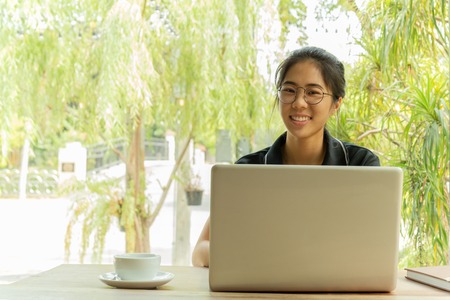 Asian woman with glasses smiling using laptop with cup of coffee on the table.の写真素材