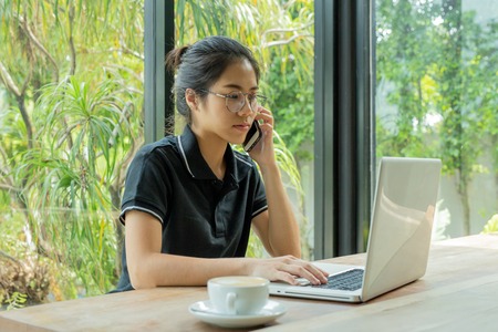 Young woman in coffee shop using a laptop and talking on a cell phone.の写真素材