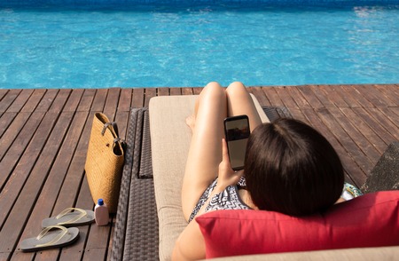 Beautiful lady using cell phone lying on chair by swimming pool with beach bag.の写真素材