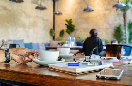 Man hand holding coffee cup with reading glasses, cell phone on table.の写真素材