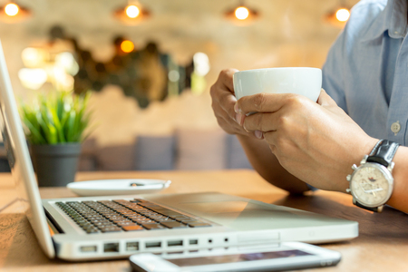Man hands holding cup coffee with mobile phone and laptop on wooden table.の写真素材
