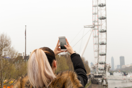 Tourist woman taking picture of London Eye with cell phone by thames river.の写真素材