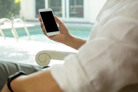 Man sit on chair near window and looking at his cell phone with swimming pool outside.の写真素材