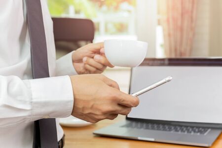 Businessmans hand holding cellphone and cup of coffee in his office.の写真素材