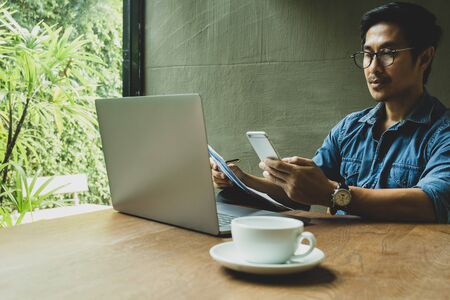 Businessman looking at cell phone and holding paperwork with laptop on table.の写真素材