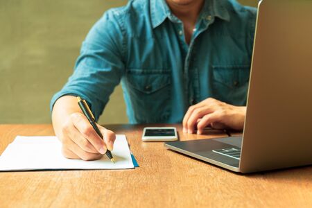 Man writing document on paperwork with laptop and cell phone on wooden desk.の写真素材