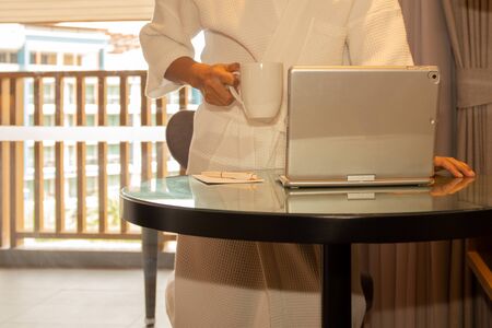 Businessman in bathrobe holding coffee mug working on laptop in hotel room.の写真素材