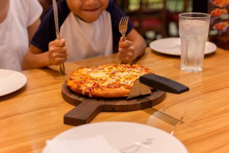 Happy little boy with hand holding knife and fork eating pizza on table.の写真素材