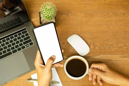 Man hand using cellphone and holding coffee cup with laptop on wooden desk.の写真素材
