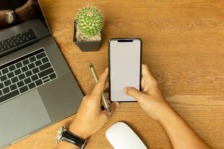 Businessman hand using cell phone and holding pen with laptop on wooden desk.の写真素材