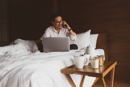 Businessman working with laptop in bed and talking on cellphone.の写真素材