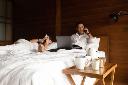 Coffee set with businessman and his wife using cell phone and laptop in bed.の写真素材