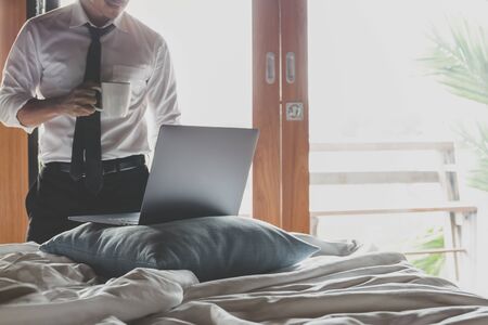Businessman holding cup of coffee and working on laptop in bed room.の写真素材