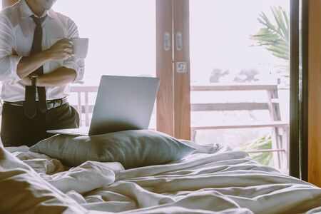 Businessman holding cup of coffee and working on laptop in bed room.の写真素材