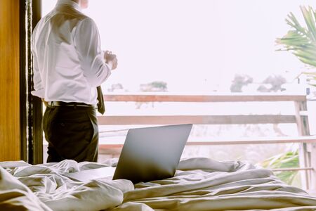 Laptop on the bed with businessman holding cup of coffee looking out hotel room.の写真素材