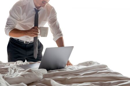 Businessman holding cup of coffee and working on laptop in bed room isolated.の写真素材