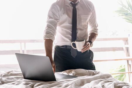 Businessman holding cup of coffee and working on laptop in hotel bed room.の写真素材