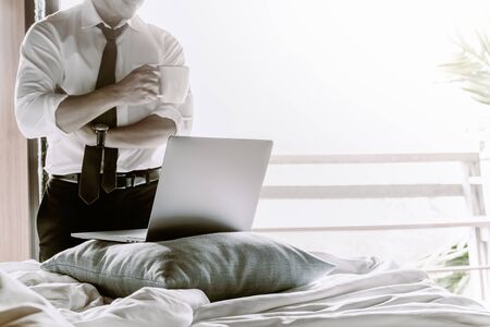 Businessman holding cup of coffee and working on laptop in bed room.の写真素材
