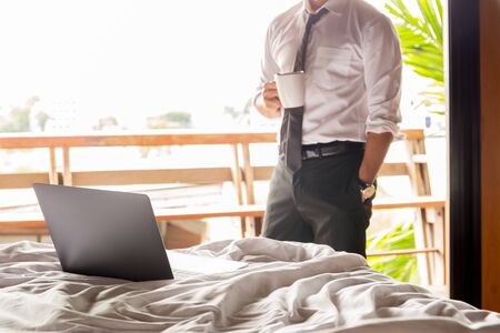 Focus laptop on the bed with businessman holding coffee in cup in background.の写真素材