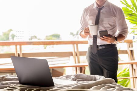 Laptop on the bed with businessman using cellphone and holding coffee in cup.の写真素材