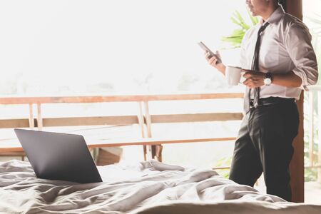 Laptop on the bed with businessman using cellphone and holding coffee in cup.の写真素材