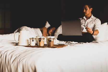 Coffee set with businessman working on laptop while his wife sleeping in bed.の写真素材