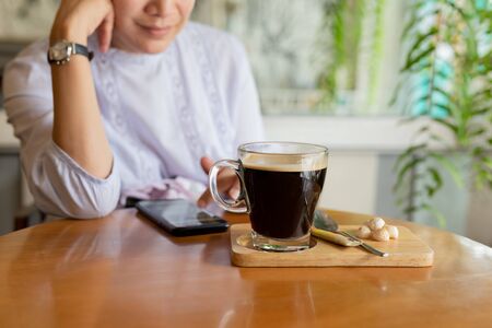 Selected focus coffee cup on table with woman using cell phone in cafe.の写真素材