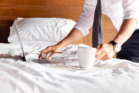 Businessman working on laptop with hand holding coffee mug in bedroom.の写真素材