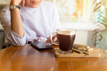 Selected focus coffee cup on table with woman using cell phone in cafe.の写真素材