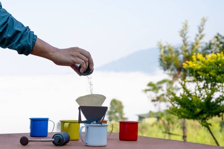 Man hand pouring ground coffee into drip coffee cup on misty mountain view.の写真素材