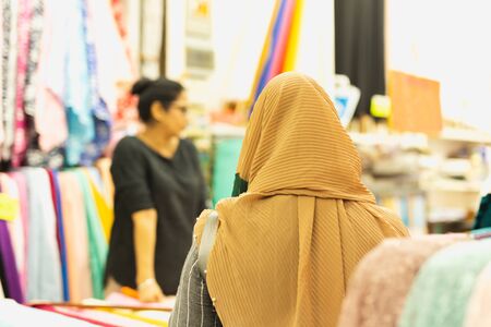 Back view of Muslim woman with cream hijab choosing fabrics at market.の写真素材