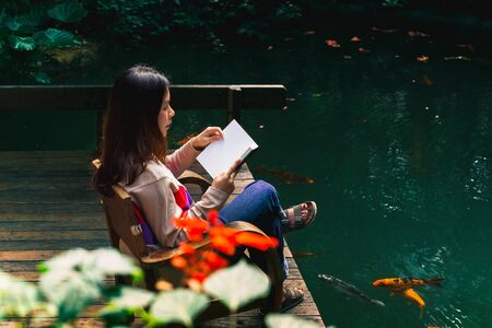 Young woman reading a book in the nature park next to the pond.