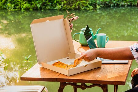 Man hand holding slice of pizza, and working on laptop computer.
