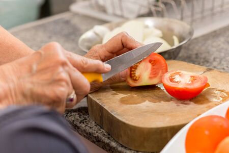 Old woman hands cutting a tomato on wooden board with knife in kitchenの写真素材