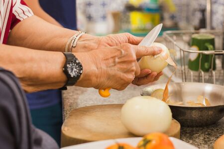 Senior woman hand peeling fresh onion with knife in kitchen.