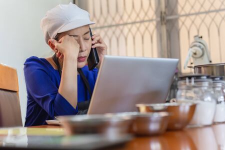 Serious woman baker talking on cell phone with laptop and bakery ingredient on table.の写真素材