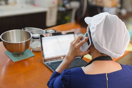Woman baker talking on cell phone with laptop and bakery ingredient on table.の写真素材