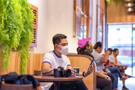 New normal man in facemask looking at smartphone with coffee and camera on table in cafe.の写真素材