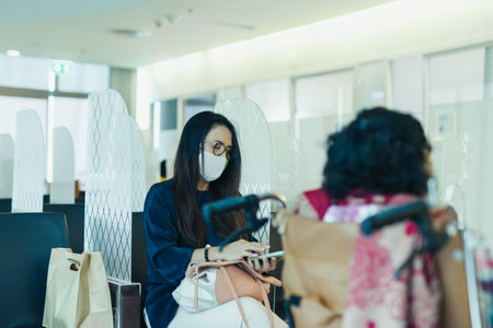 Woman in protective mask using cell phone sitting in partition with her senior mother in hospital.の写真素材