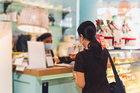Woman with protective mask paying bill at cashier counter in cafe.