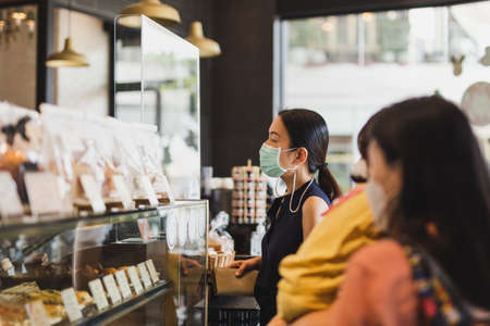 Social distancing women in medical mask ordering dring at cafe counter with partition.