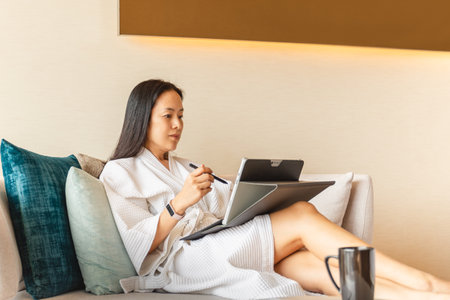 Woman in dressing gown sit on couch in hotel room working on laptop.の写真素材
