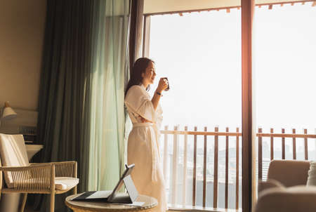 Happy businesswoman in dressing gown stands near the window holding cup of coffee.の写真素材