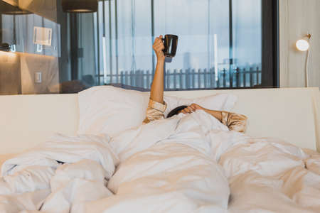 woman hands holding cup of coffee above blanket in bed.の写真素材