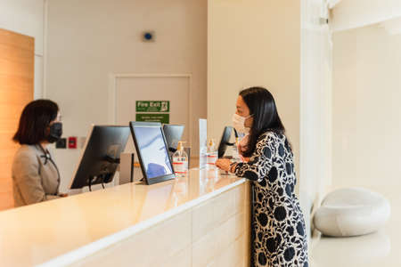 Woman hotel guest and receptionist at hotel counter wearing medical mask against virus.の写真素材