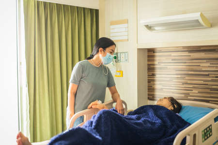 Daughter in medical mask visiting her mother lying in bed at hospital ward.の写真素材