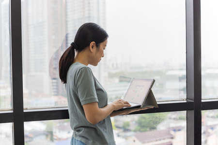 Woman standing next to window holding laptop computer.