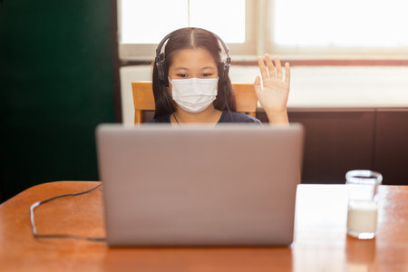 Asian student in face mask waving hand while holding class over a laptop duringの写真素材